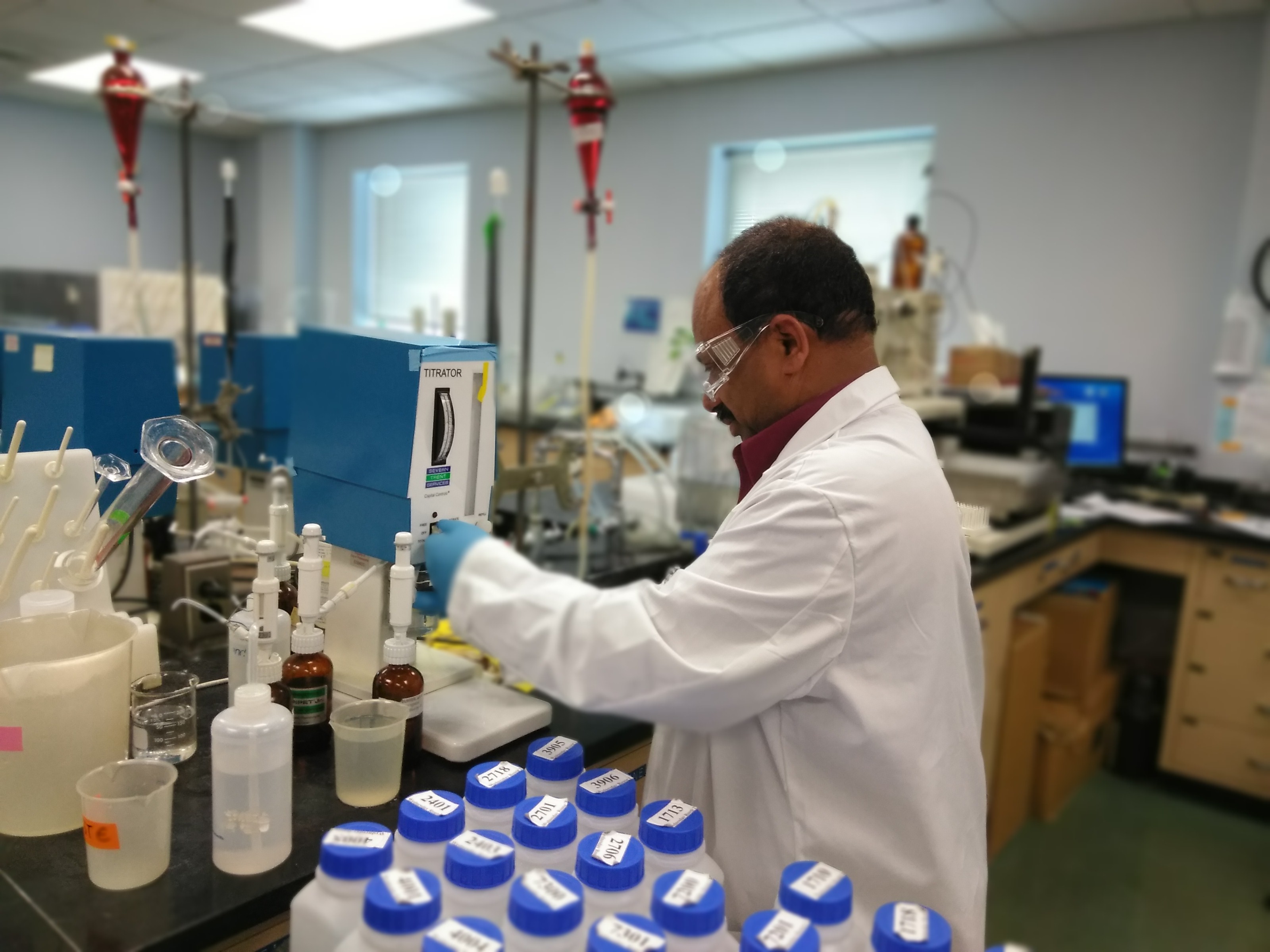 A photo of a scientist in a white lab coat, rubber gloves, and safety glasses uses a machine to test a water sample.