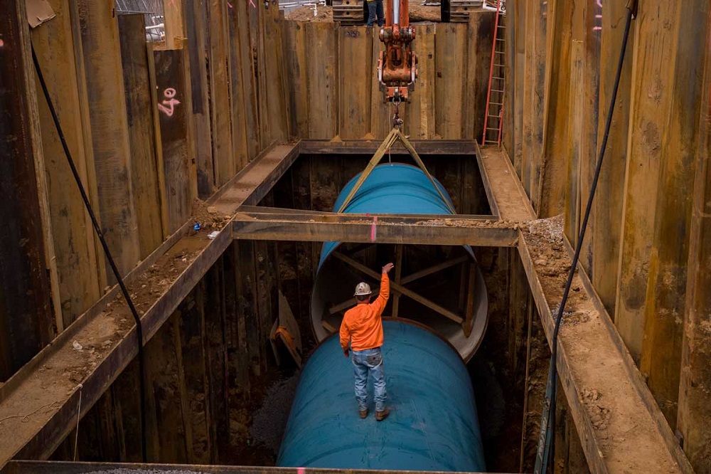 Water mains 10-feet in diameter are being installed underground. A man in a hardhat stands in a pit on one of the mains and helps direct a crane lowering another 10-foot pipe to connect to the other segment.