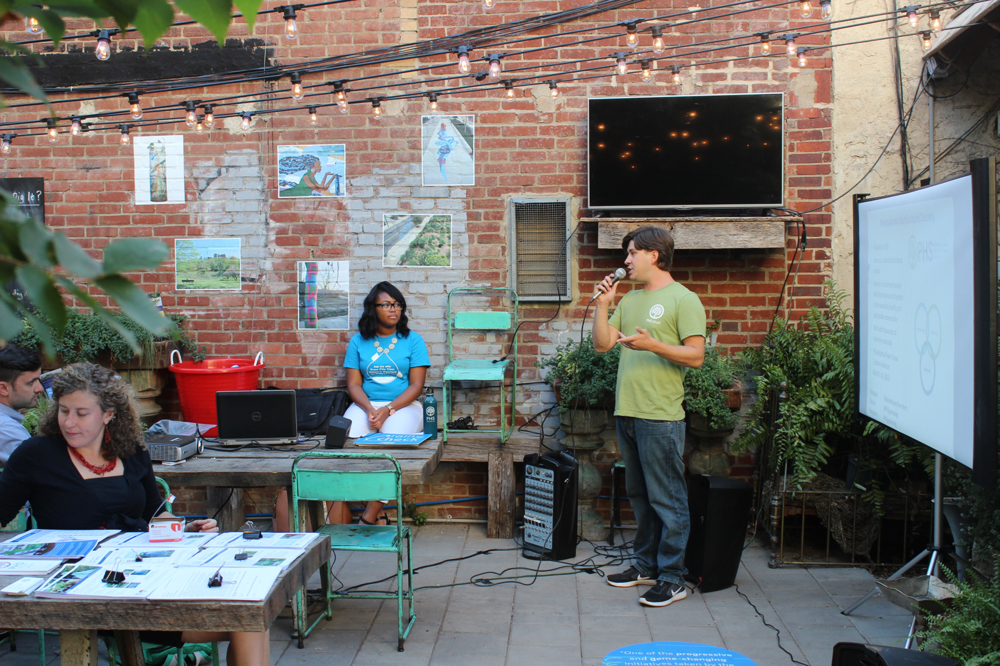 A photo from a past workshop shows a presenter speaking into a microphone, explaining a diagram projected onto a portable screen. Workshop participants are seated at large wooden tables in a brick-walled courtyard with strands of lights strung back and forth overhead and lots of potted plants around the edges.