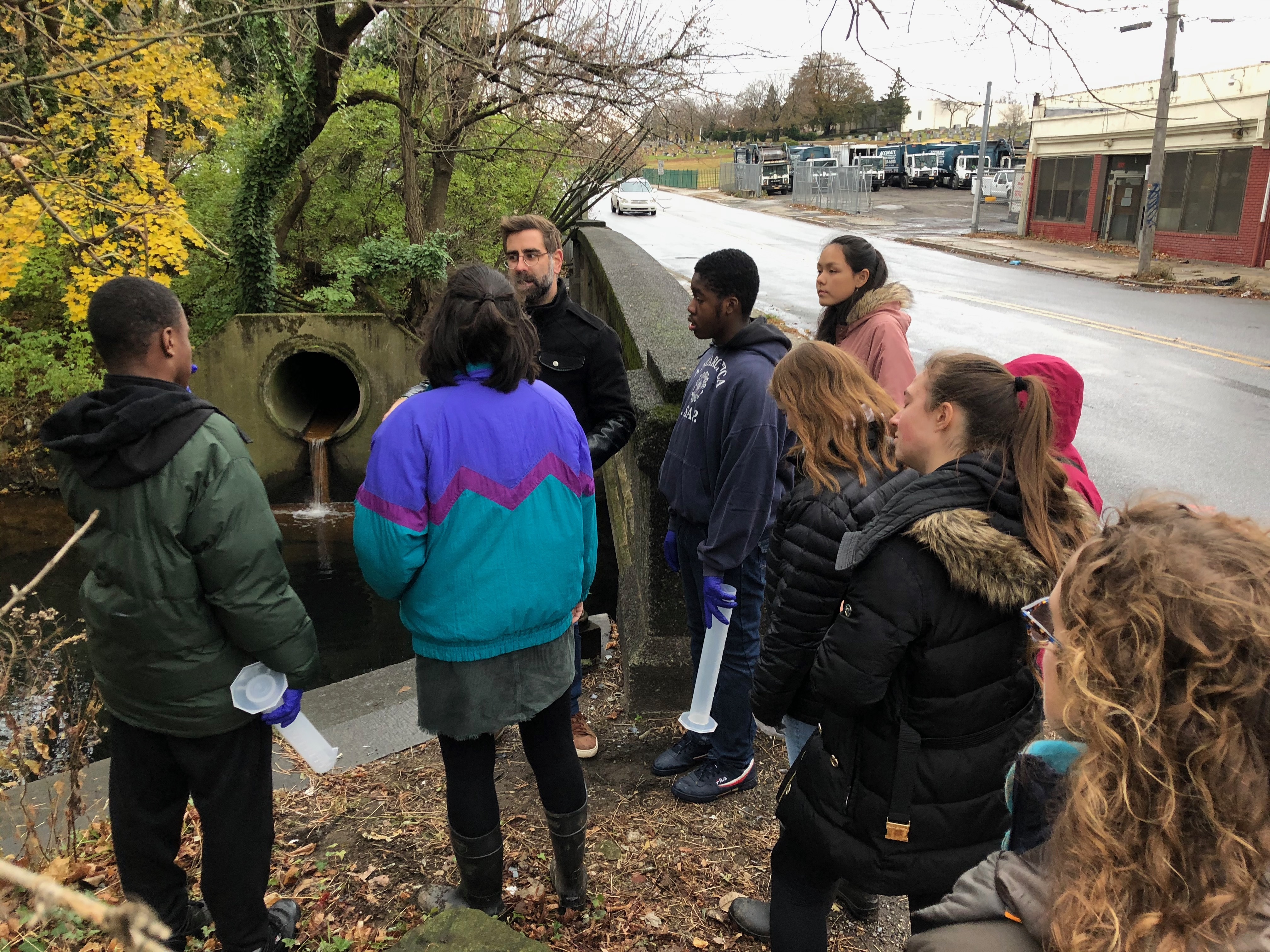 PWD Outreach Specialist Dan Schupsky leads a tour group on a rainy day, as a an outfall in the background trickles a small amount of runoff into a creek