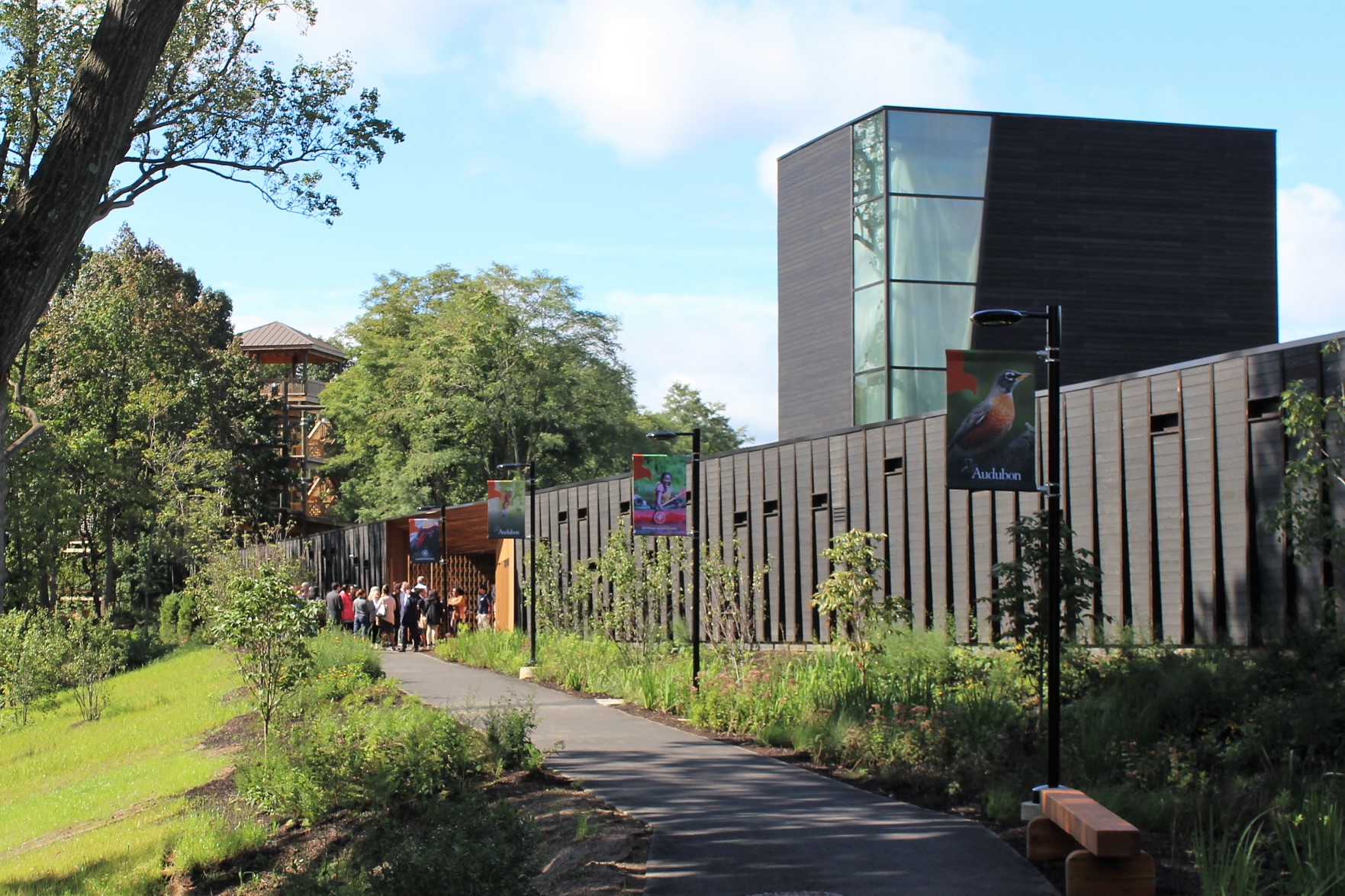A photograph of the entrance to the new Discovery Center, shows off its modern architecture. Looking down the path lined with greenery and lampposts bearing flags featuring Audobon and Outward Bound activities and nature scenes, a crowd is gathered at the cedar-lined pass-through to the reservoir. In the background, a structure from the Outward Bound High Ropes Course can be seen through a gap in the trees.