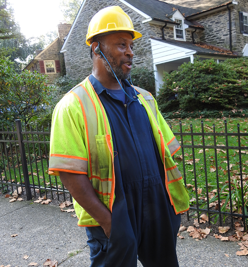 Lance Johnson, heavy equipment operator, is wearing a hard hat and safety vest and smiles as he talks. The big homes of East Falls are made of stone. 