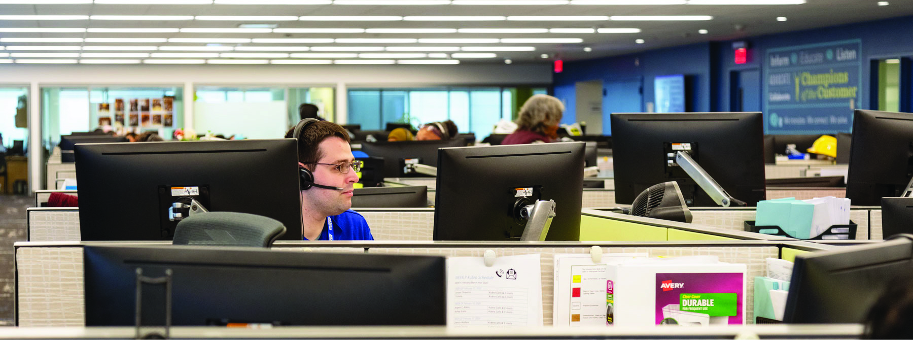 A glimpse into the Philadelphia Water Department Contact Center: pairs of computer monitors on movable arms peek above low cubicle walls. In one of the nearest cubes, a representative with short brown hair, glasses, a bit of a five-o-clock shadow, and a headset looks at a monitor. Others are partially visible, though out of focus, in the distance, against a backdrop of glass-walled offices and a conference room, and one side wall painted dark blue.