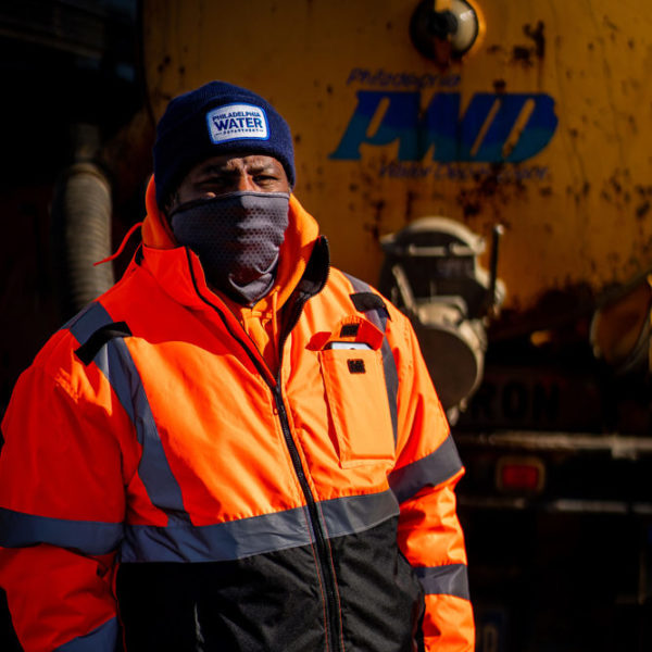A man wearing a high-visibility orange coat, navy blue winter cap with the Philadelphia Water Department logo, and grey gaiter pulled up over his mouth and nose stands in front of a yellow vehicle with the old PWD logo on the side.