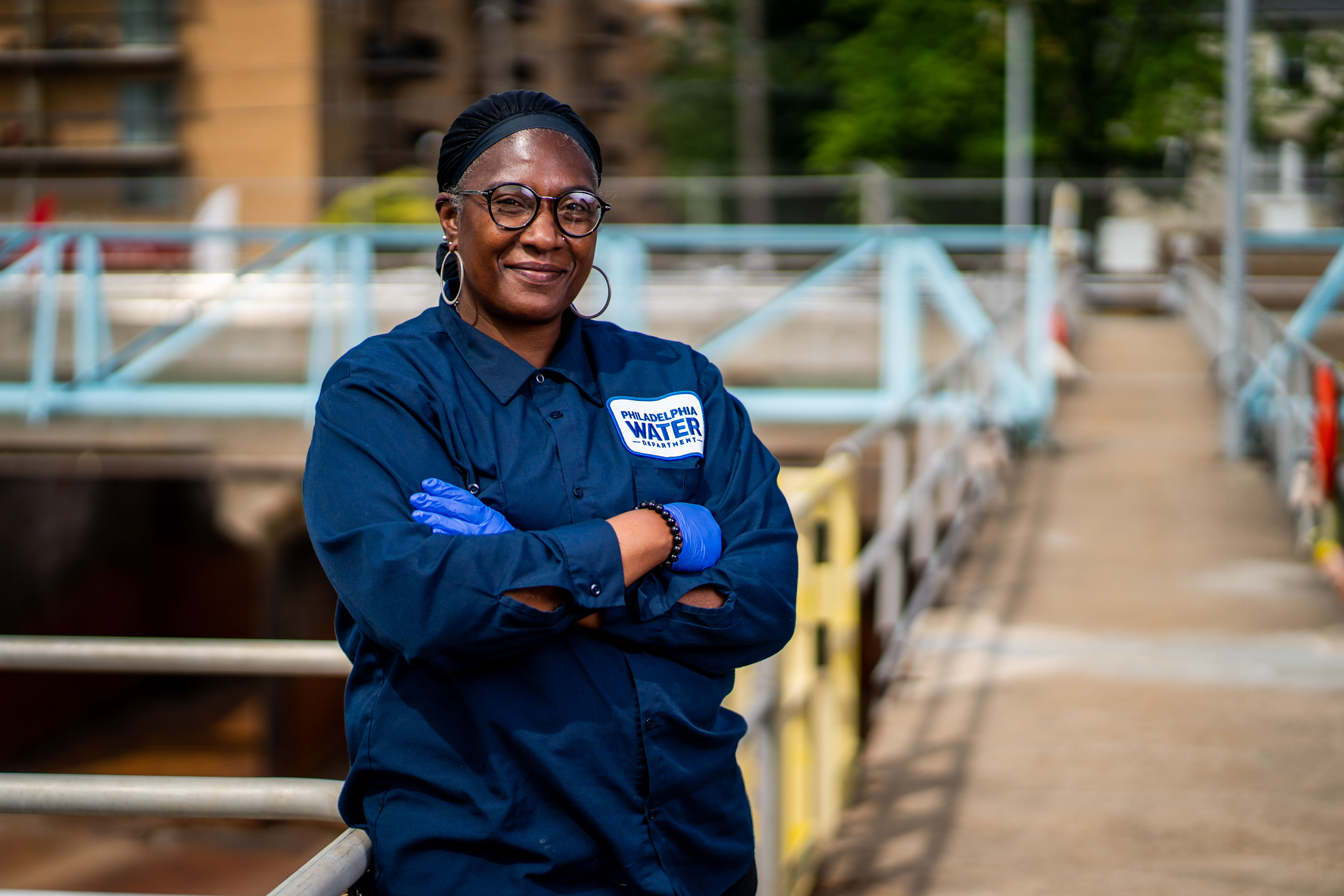 A smiling PWD employee stands with arms crossed at one of our water treatment plants, wearing navy blue coveralls with a "Philadelphia Water" patch, black glasses, hoop earrings, a black head wrap, and blue disposable gloves.