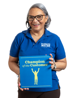 A smiling PWD Contact Center representative with chin-length straight silver hair, wearing a headset, black glasses, a blue PWD polo shirt, and black slacks holds up a card that reads, "Champion of the Customer", with a silhouette of a person standing with arms raised.