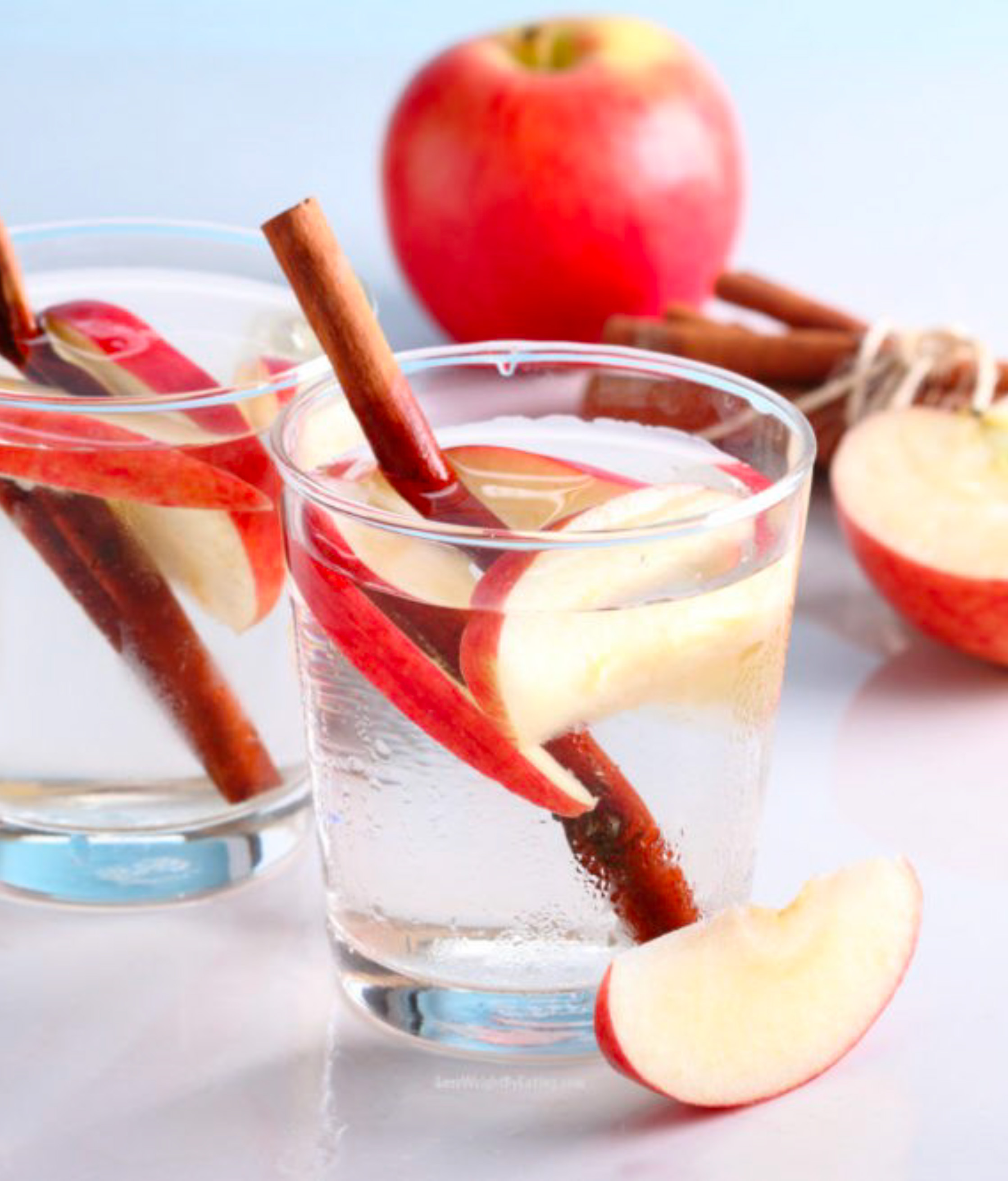 Glasses of water with a cinnamon stick and several thin slices of red apple in each, sitting on a white surface, one centered while the other sits off to the side a little behind it, with another slice in front of the centered glass, and a whole apple, a partially sliced apple, and more cinnamon sticks out of focus in the background