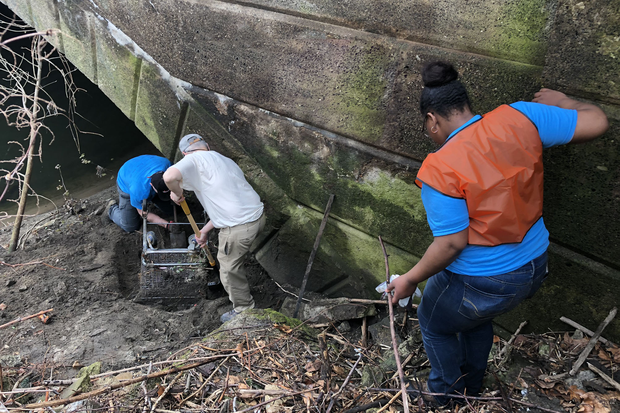 Two are at the bottom of the hill using shovels to excavate an overturned shopping cart partially buried in the silt at the edge of a creek, under the arc of a concrete bridge. A third, holding a pair of work gloves in one hand while bracing against the bridge with the other, carefully steps down the hill to join them.