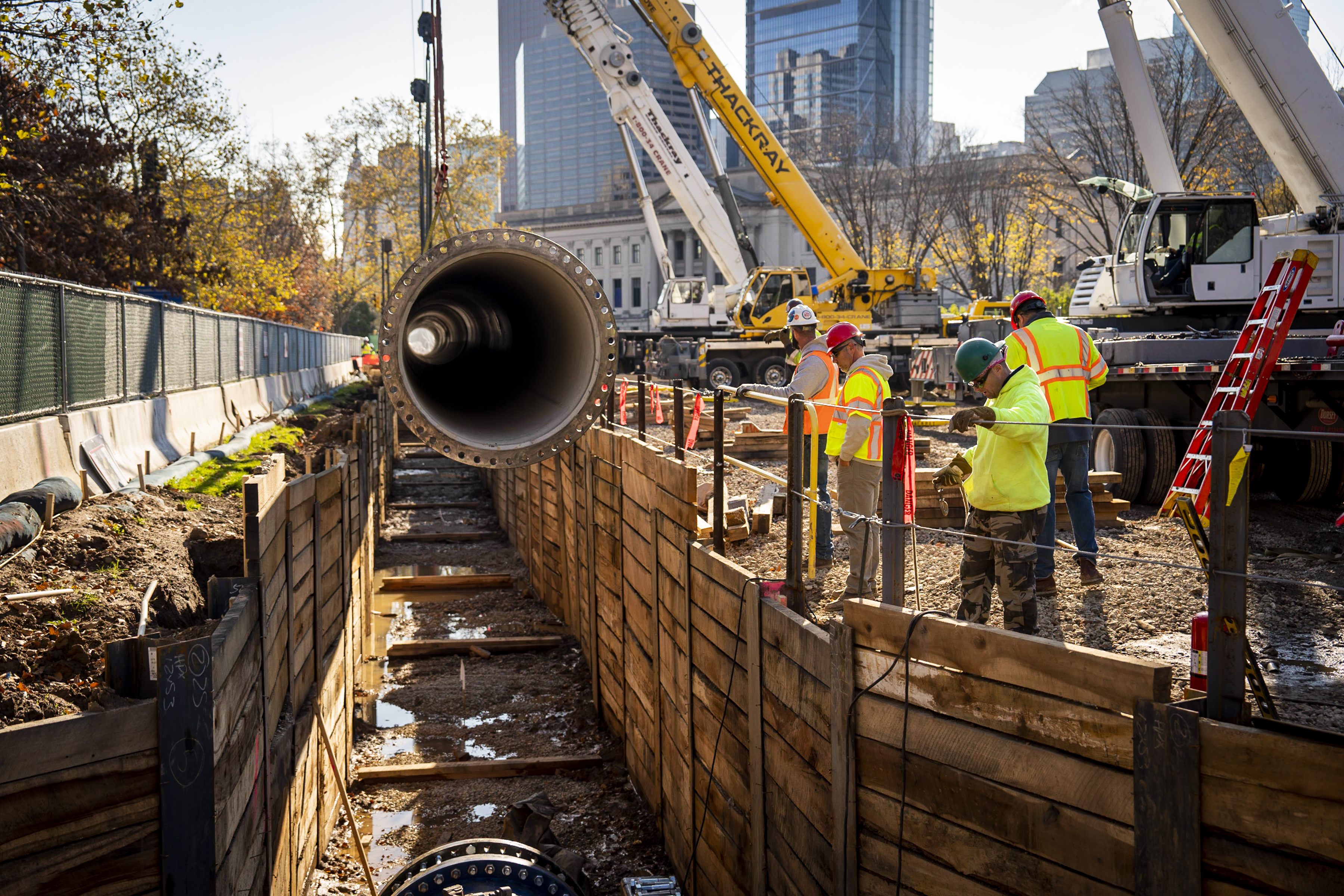A section of new water main is lowered into a trench stabilized with wood plank walls holding up the sides. We're looking down the length of the trench and into one end of the pipe, which has a raised ring on the end, with holes for bolting it onto the next section all the way around it. Two crane arms are visible in the top right corner, and behind them we see a bit of the Franklin Institute, One and Three Logan Square, and other buildings in the background. Trees with fall foliage line the left side, behind the construction fencing.