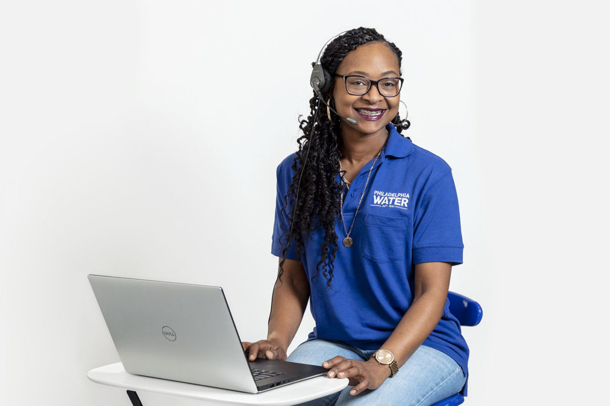 A smiling PWD contact center representative with long, curly, black hair twisted into spirals, wearing a headset, blue PWD polo shirt, and jeans, with black glasses and gold jewelry, sits at a small table with hands resting lightly on the edges of a laptop computer.