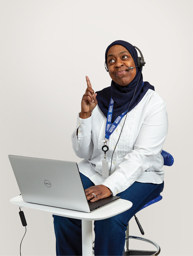 A PWD Customer Service representative is shown seated on a stool at a small desk with a laptop on it. She is wearing jeans, a white long-sleeved shirt, black hijab, headset, and blue PWD lanyard. She has a knowing look on her face as she points to the text, "Your account balance will still grow with every unpaid bill."
