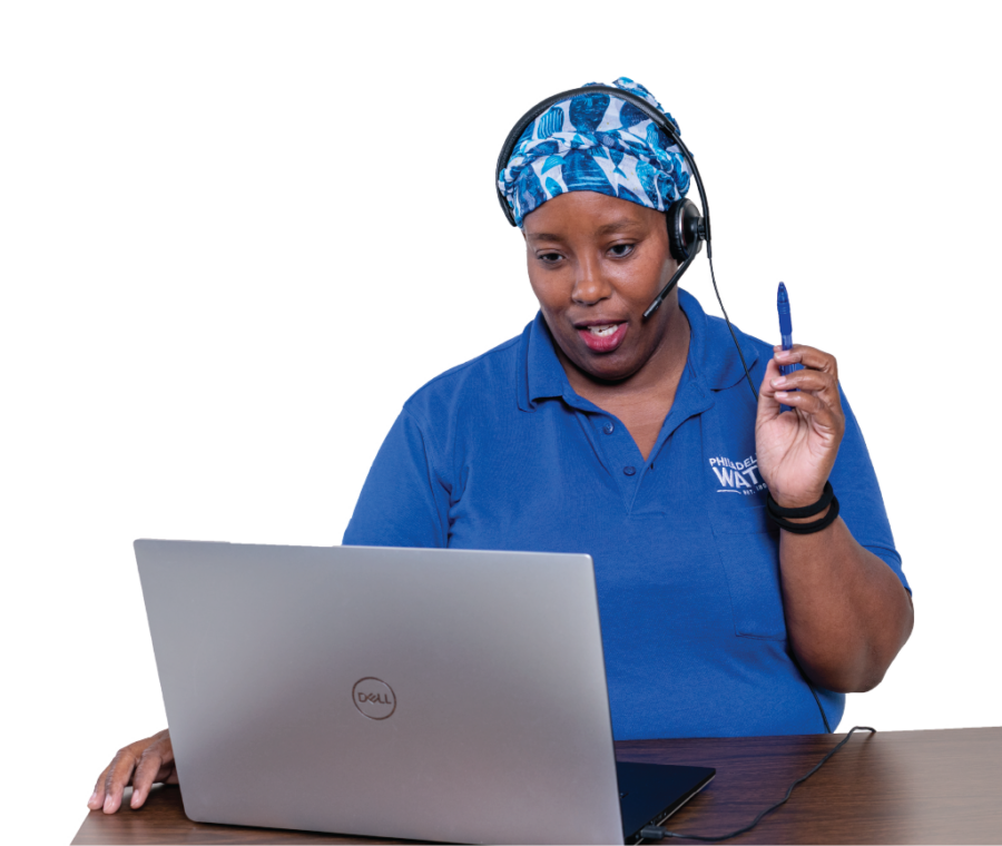 A contact center representative wearing a blue PWD polo, a headwrap with a vibrant blue and white fish pattern, two black bracelets, and a phone headset sits at a desk, looking at a laptop, and seems to be speaking with a customer, mouth partially open and a pen held in the air as if in thought.