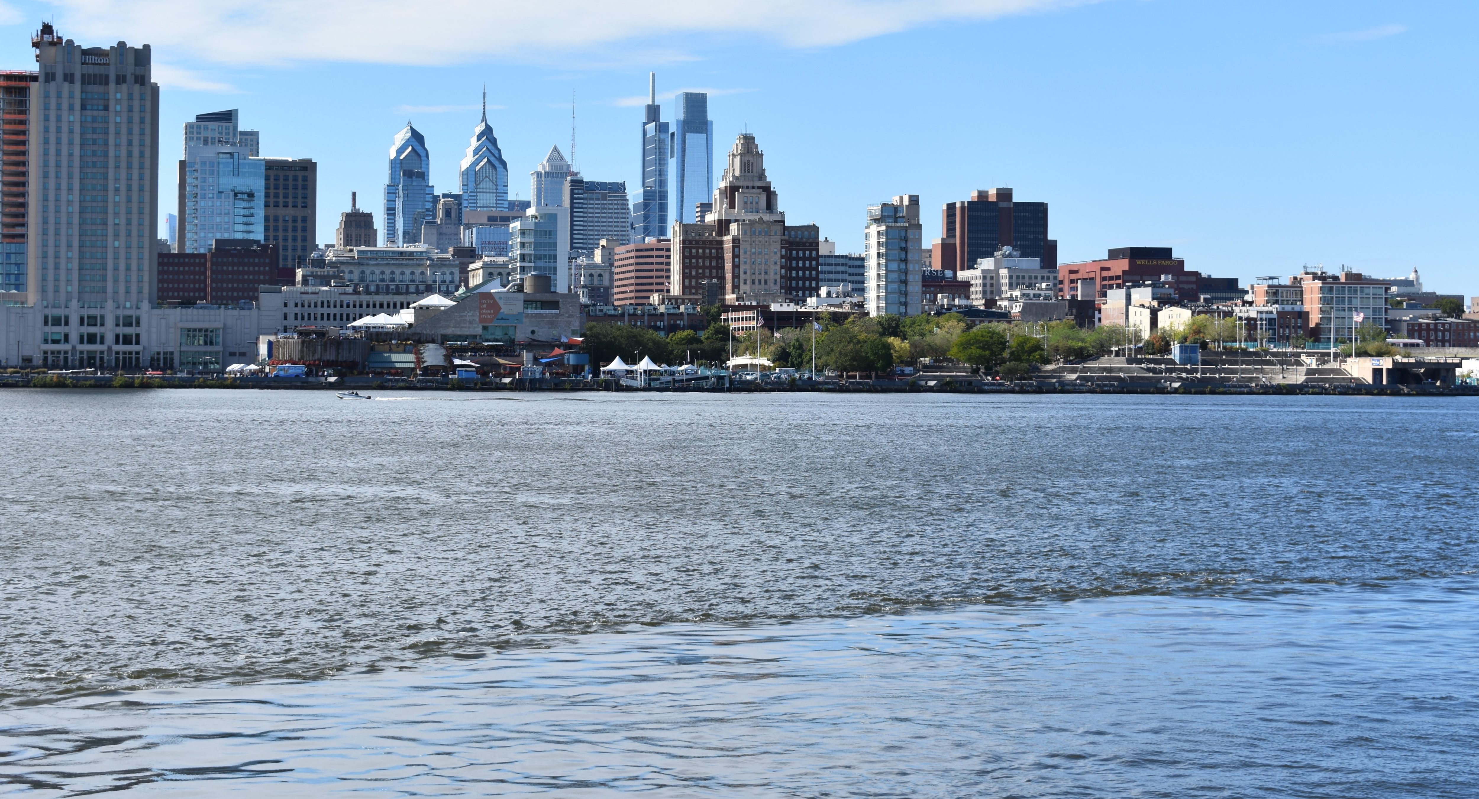 Looking out over the Delaware River from Camden, NJ, on a beautiful day, the Philadelphia skyline shines between a mostly blue sky and the shimmering river. The tents, crowds, and an ice cream truck for Delaware River Fest 2022 are visible on Penn's Landing.