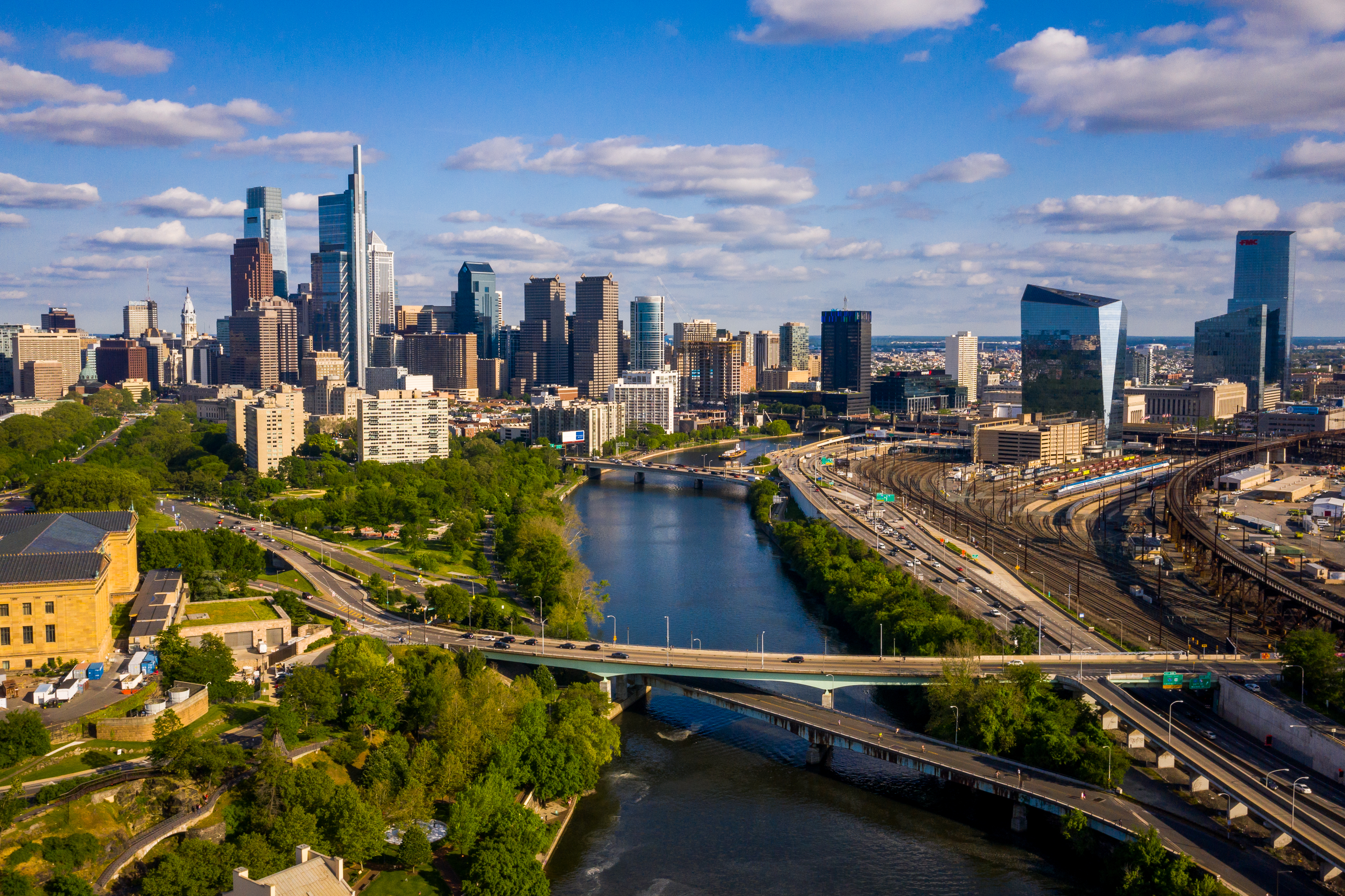 an aerial photo taken over the Schuylkill river near the Fairmount Water Works, looking out over the river downstream towards Center City Philadelphia, with the Art Museum partially visible on the left, 30th St. Station on the right, and bits of South Philly and the Delaware River in the background.
