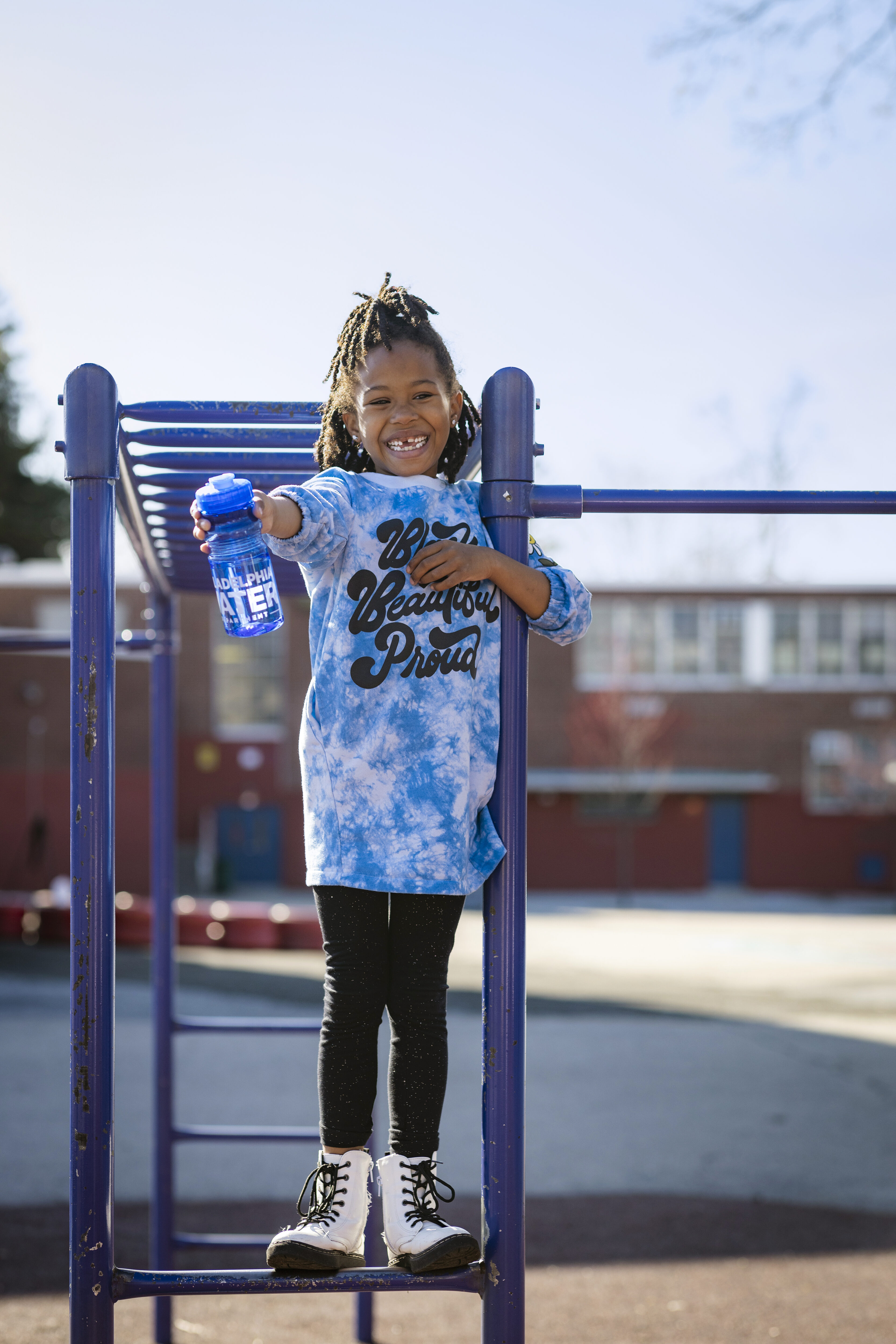 A young girl with a huge smile showing off a missing tooth as she perches on the monkey bars at Ellwood Elementary School, holding up her reusable PWD water bottle. She has brown skin, dark hair in shoulder length braids, half pulled up on top of her head, and is wearing an oversized blue tie-dye tshirt with black leggings and white lace-up boots.