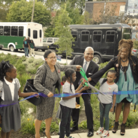 a row of PWD employees, community members, and children hold a ribbon stretched out in front of a newly completed rain garden, with several helping to hold very large scissors, poised to cut it