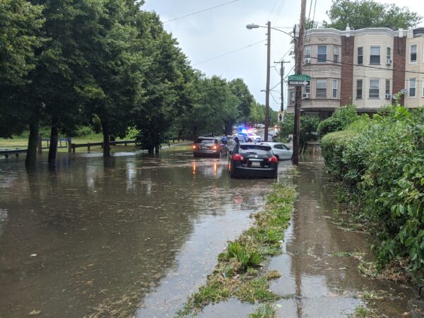 flooding at the intersection of E. Haines St. and Crittenden St. fills covers the street, sidewalks on both sides, and spills into the parkland at Awbury Recreation Center. A police car blocks the road in the background as an officer approaches a stranded vehicle. Other cars seem to have been lifted from their parking places by the rising water.