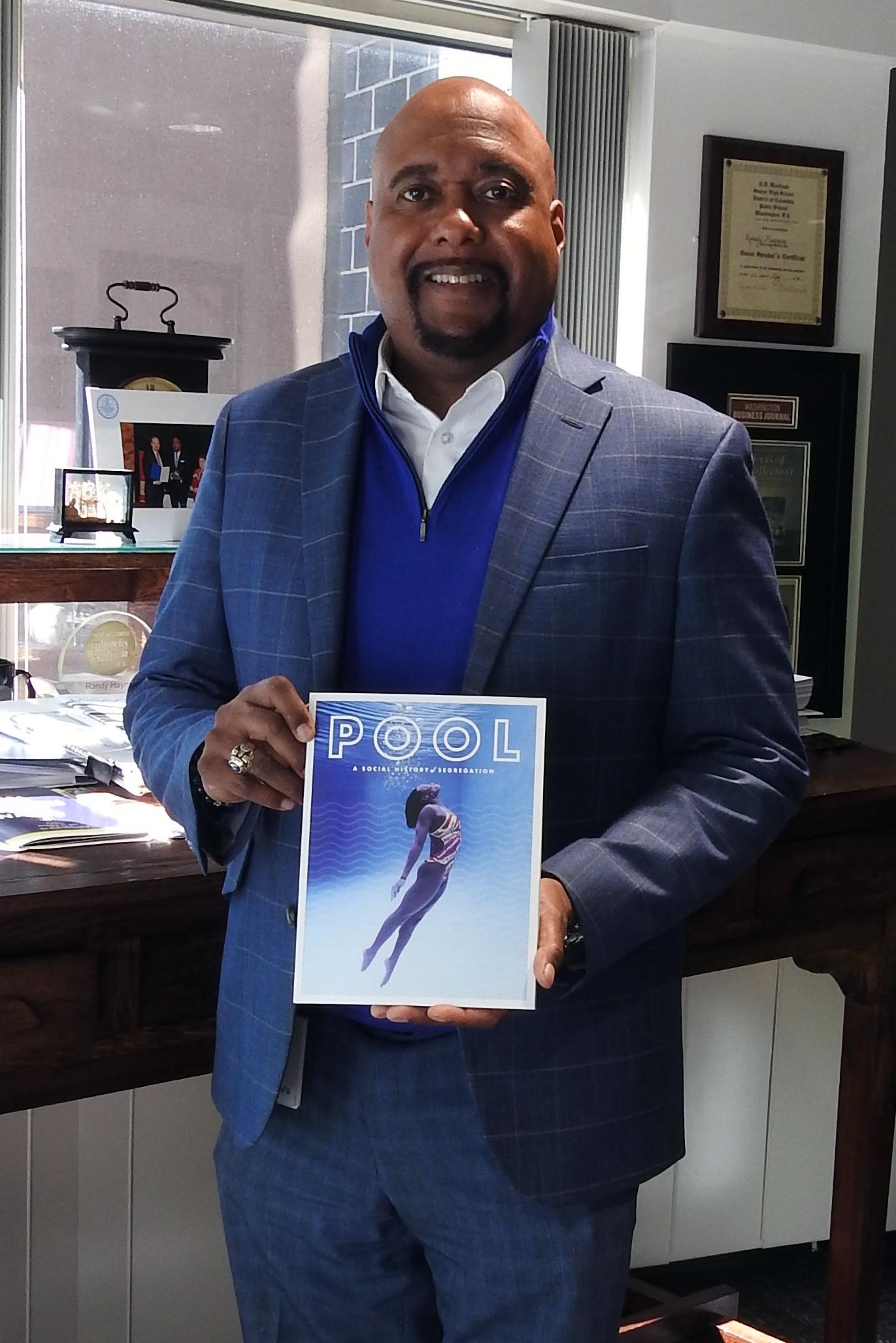 PWD Commissioner Randy Hayman, a black man with a clean-shaven head and black goatee, wearing a crisp white shirt under a cobalt half-zip sweater and medium blue suit, stands in front of an antique wooden desk in his office, displaying a printed copy of the POOL magazine, which features an underwater shot of a young black girl in a swimsuit rising up from the bottom of a deep pool, surrounded by bubbles, and the word POOL outlined in white across the top.