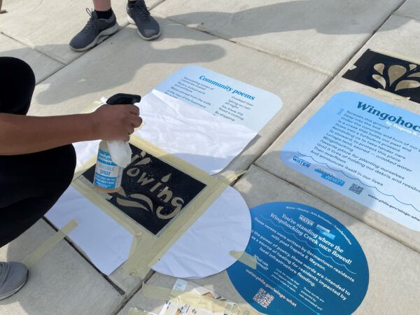 PWD staff stenciling the sidewalk with water-activated "invisible" paint (to show water-themed words and graphics on the pavement when it rains), next to decals featuring information and poems from the Wingo-WHAT? project