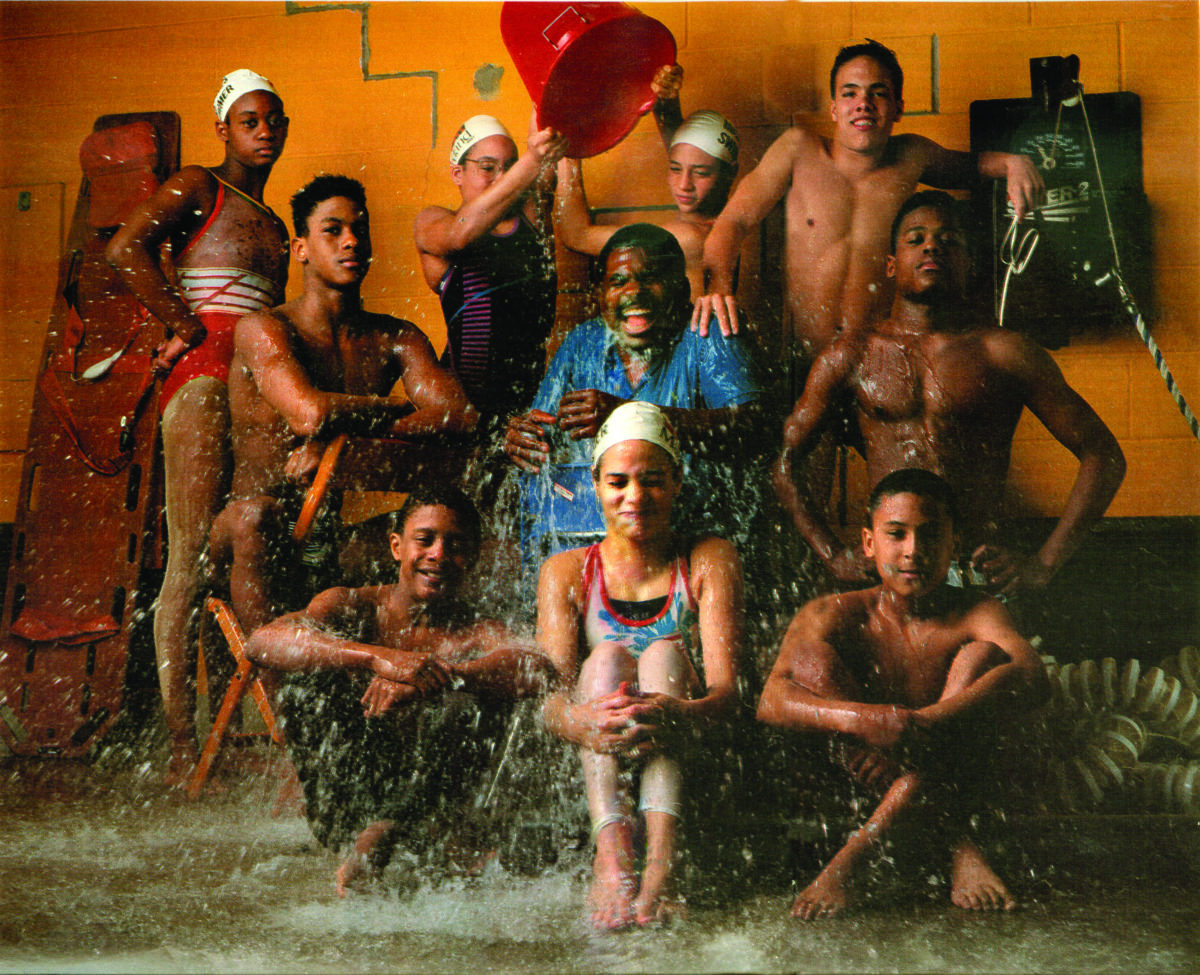Jim Ellis, with brown skin, short black hair and mustache, wearing a blue short sleeved shirt, sits in the middle of a diverse group of young swimmers posed for a photo in swim suits and some in swim caps, laughing as two in the back dump a large bucket of water over his head, splashing onto the kids in the front row and all over.