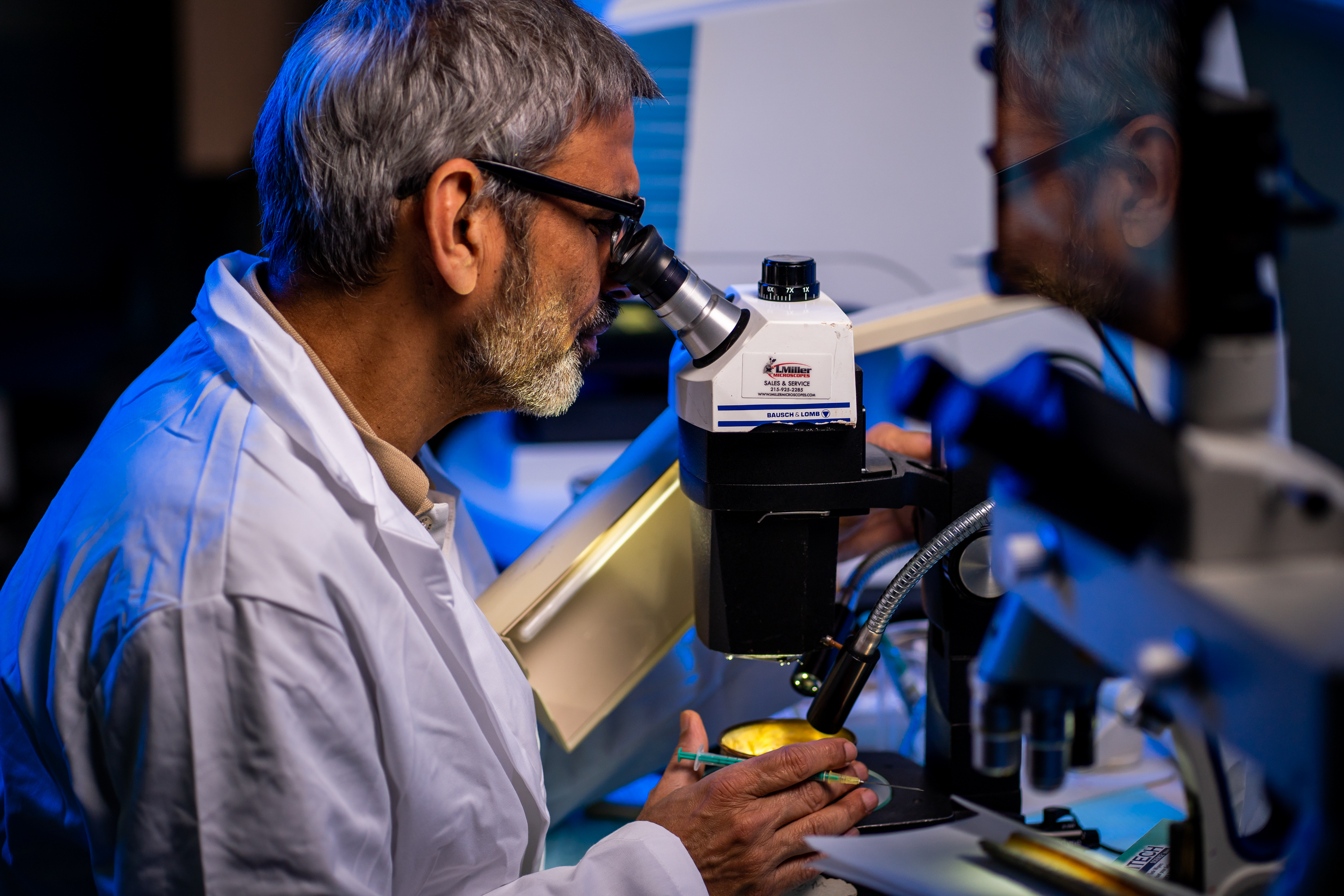 A scientist with short salt and pepper hair and close cropped beard, wearing a white lab coat and black glasses is seen in profile, peering into a microscope with pencil in hand.