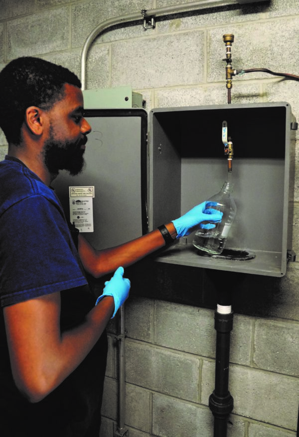 photo of a man filling a glass bottle from a monitoring tap in a utility box mounted on a cinderblock wall.