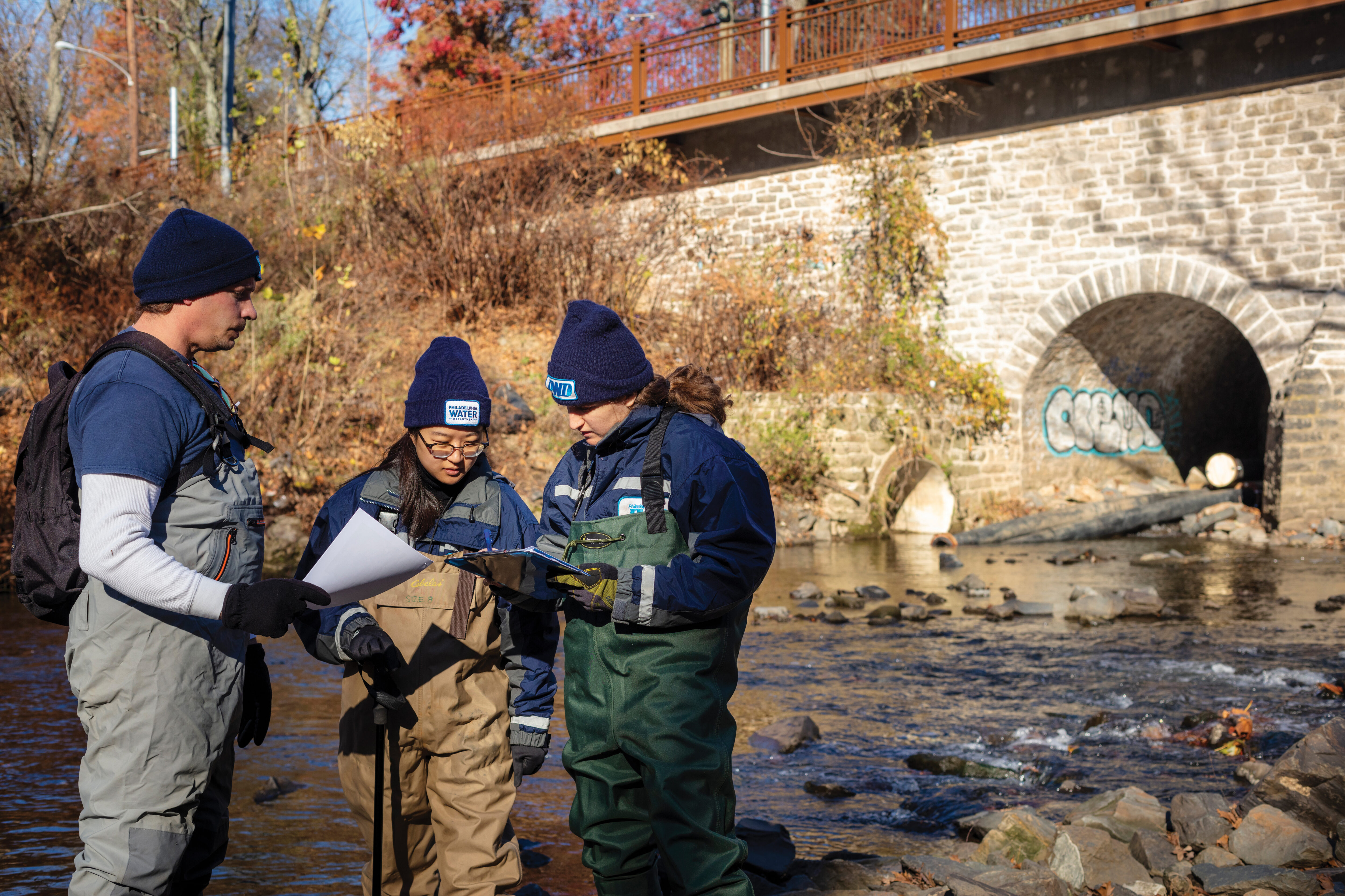 Three PWD employees in dark blue shirts or jackets and winter hats with PWD logos on them, and chest-high waders in different colors, stand in Pennypack Creek, in front of an arched stone bridge to their right and fall foliage on the opposite bank seen to the left, looking at a clipboard during an assessment of the stream.