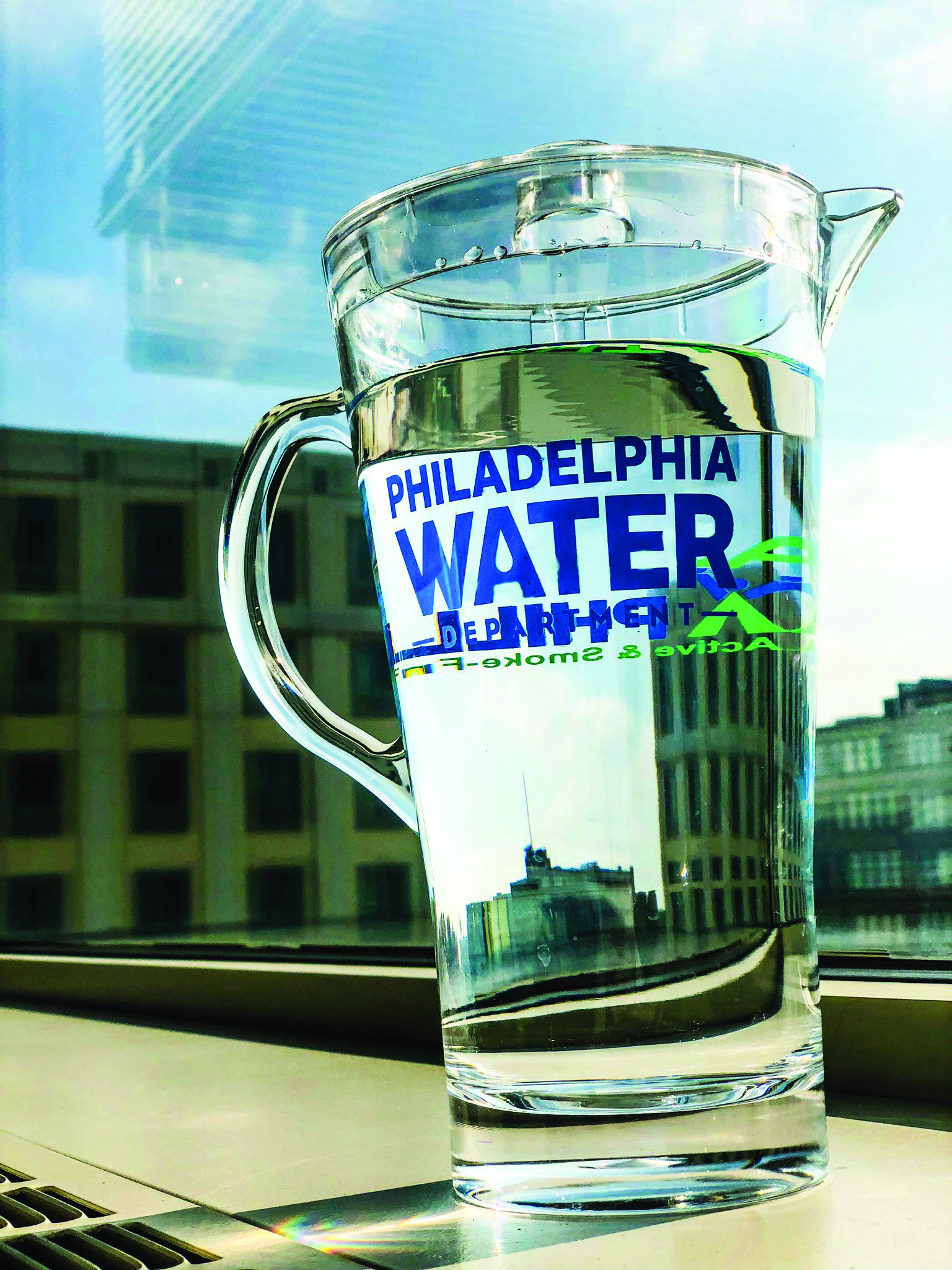 a clear plastic pitcher with the Philadelphia Water Department logo on it, full of water, sitting on a windowsill with city buildings and a blue sky visible in the background