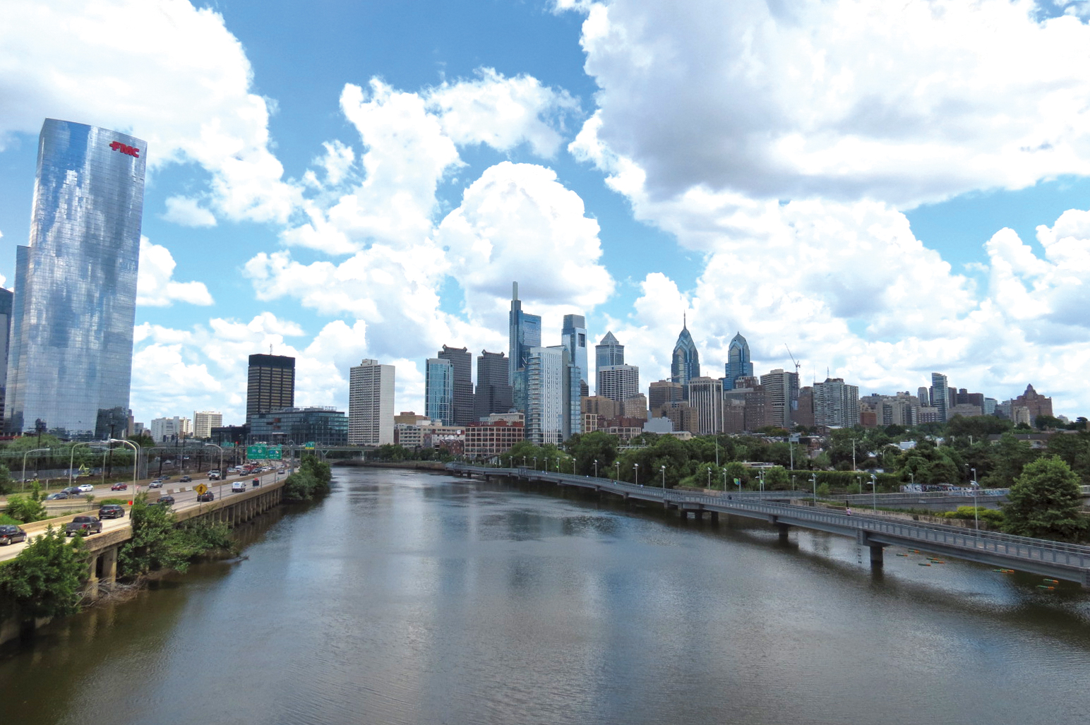 A view taken over the Schuylkill River, looking upstream, from around the South Street Bridge. As the river curves off to the left, the Center City skyline is front and center. The Schuylkill Boardwalk is visible along the right side of the river, I-76 Schuylkill Expressway on the left, and the top half of the picture is dominated by a blue sky full of fluffy white clouds.