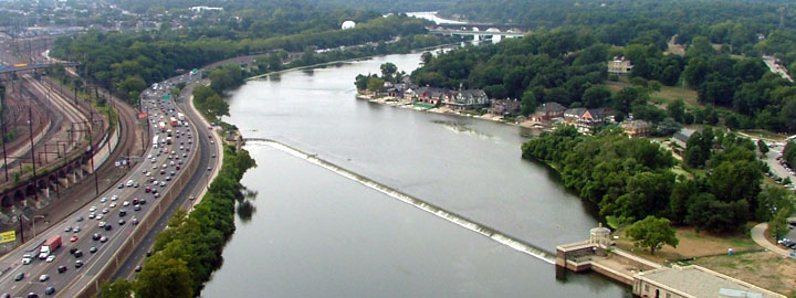 Color aerial of a river, with a freeway, railroad tracks, a dam, houses and trees visible. 