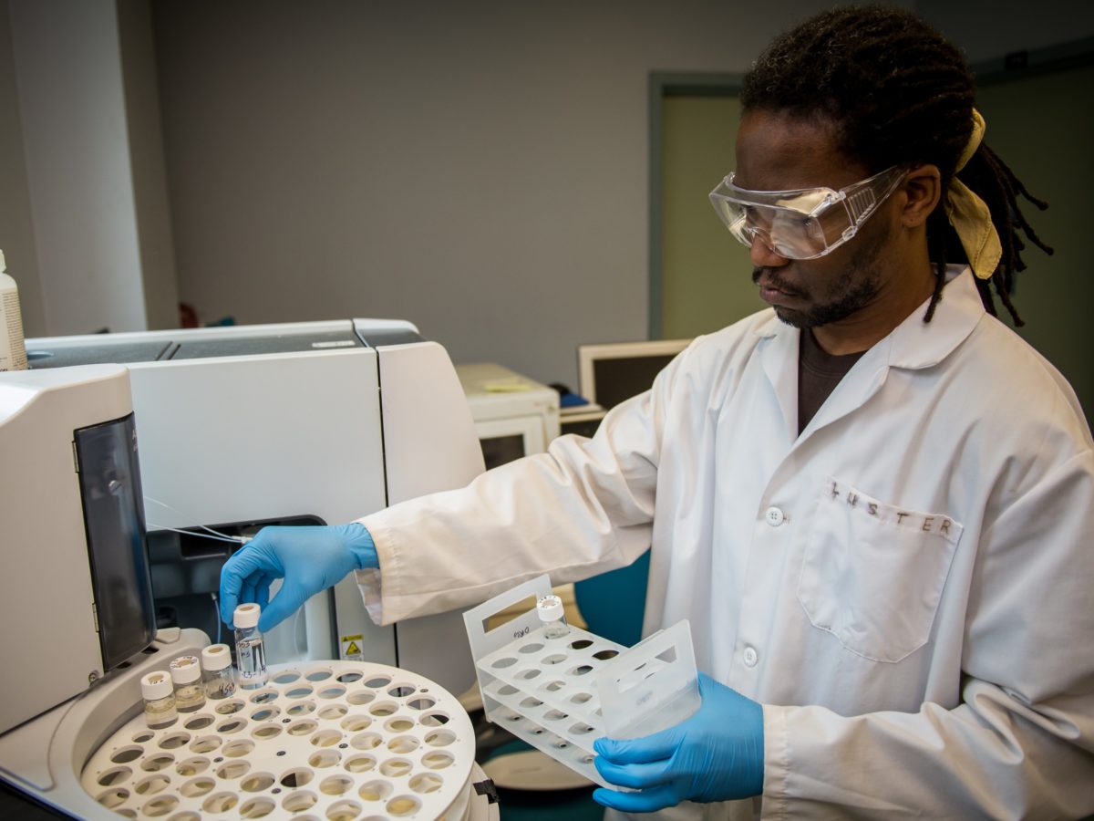 Lerone, with his neat shoulder-length locs tied back and short black facial hair, wears safety goggles, a white lab coat, and blue nitrile gloves, as he places water samples in glass vials into a circular rack with four concentric rings of round holes at the front of a piece of equipment.