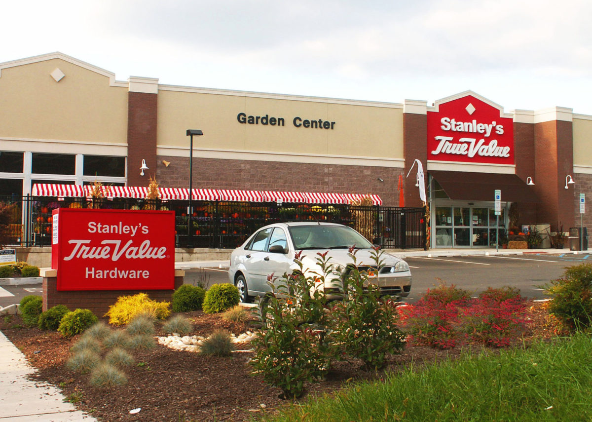 Stanley's True Value Hardware has a large storefront with light reddish stone on the bottom and beige stucco on the upper portion of the facade, and columns of red brick framing the entrance, a red and white awning in front of the Garden Center, and signs with white lettering on a bright red background above the entrance and in the rain garden, which is visible in the foreground. A car in the parking lot is partially obscured by plants in the rain garden.