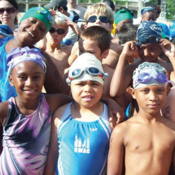 a diverse group of young children in swim suits, many wearing swim caps and goggles or sunglasses, stand close together for the picture, some smiling for the camera, others squinting at the bright sunlight.