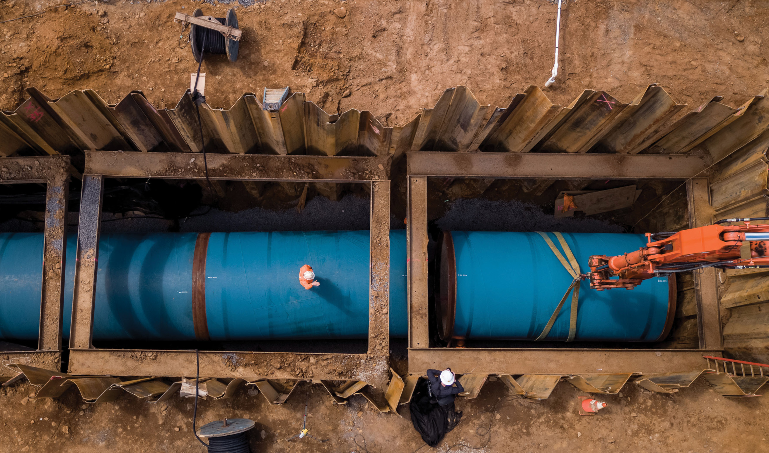 Arial view of a construction site where a section of water transmission main is being laid. We're looking down into a large trench, dug into and surrounded by reddish dirt, with the sides supported by massive corrugated metal panels and steel beams as an orange crane arm lowers a piece of bright blue pipe, at least 10 feet wide and 20 long, into the hole. Workers can be seen looking on, standing on another section of the pipe and on the side of the trench, and seem very small.
