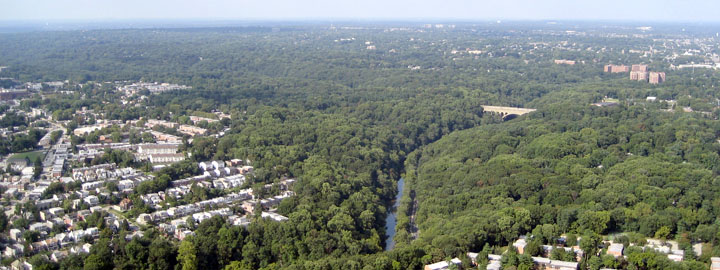 Color aerial shot of a stream flowing between two heavily wooded banks, with neighborhoods visible on either side
