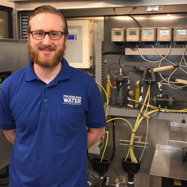 A PWD employee with reddish brown slicked-back hair and short beard, wearing black glasses, a blue PWD polo shirt, and blue jeans, stands smiling in front of a bank of machinery with lots of tubes, sensors, and small display panels