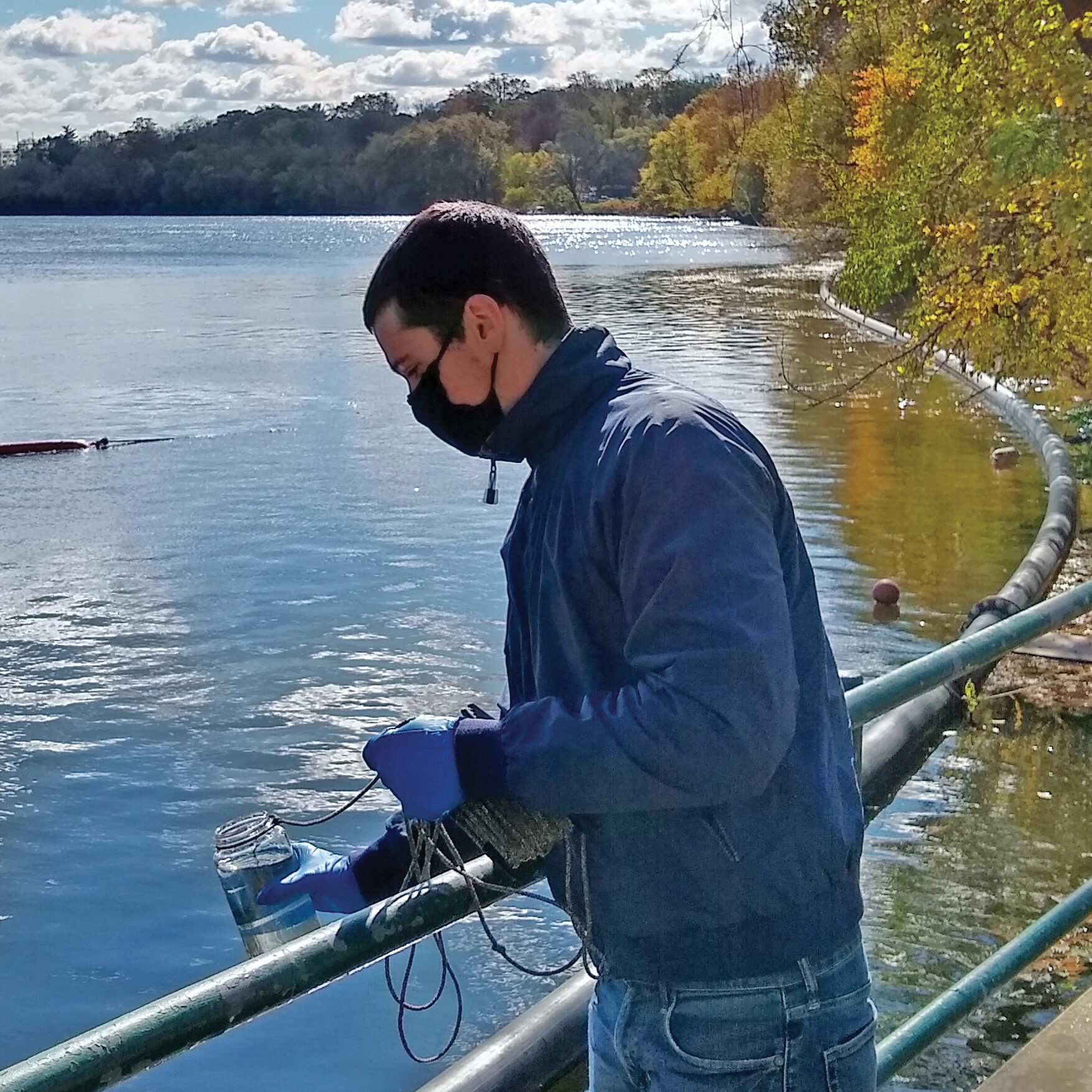 A PWD employee with short dark hair, light skin, a black face mask, blue jacket and jeans, and purple disposable gloves leans slightly over a railing on a low bridge, having holding a jar full of water attached to a cord used to lower it into the water to collect a sample for testing.