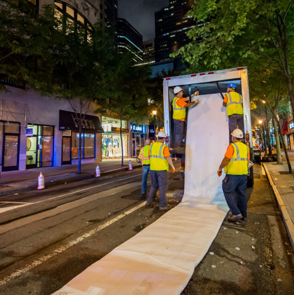 At night in Center City, a crew of 5 wearing hard hats and yellow reflective vests over orange t-shirts and blue jeans, with white and orange work gloves, unspools a section of sewer lining from the back of a box truck, laying it out flat on the street. Shops and trees line both sides of the street, and the One Liberty Place tower is visible in the background with its crown lights lit up red and the spire white.