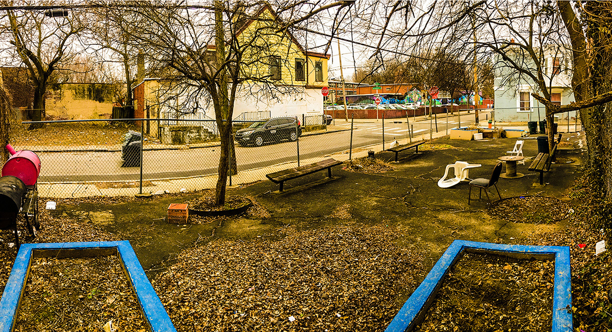 a wide angle photograph of Bringhurst Park, which is a small paved lot surrounded by a chain-link fence. There are empty raised garden beds at both ends of the park and a few trees and benches in the middle, as well as chairs and makeshift tables and a lot of fallen leaves.
