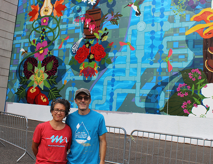 Jones and McShane are pictured standing smiling in paint-splattered t-shirts in front of their newly completed mural, full of boldly colored flora and fauna on a backdrop of waterways, pipes, and abstract water-themed motifs.