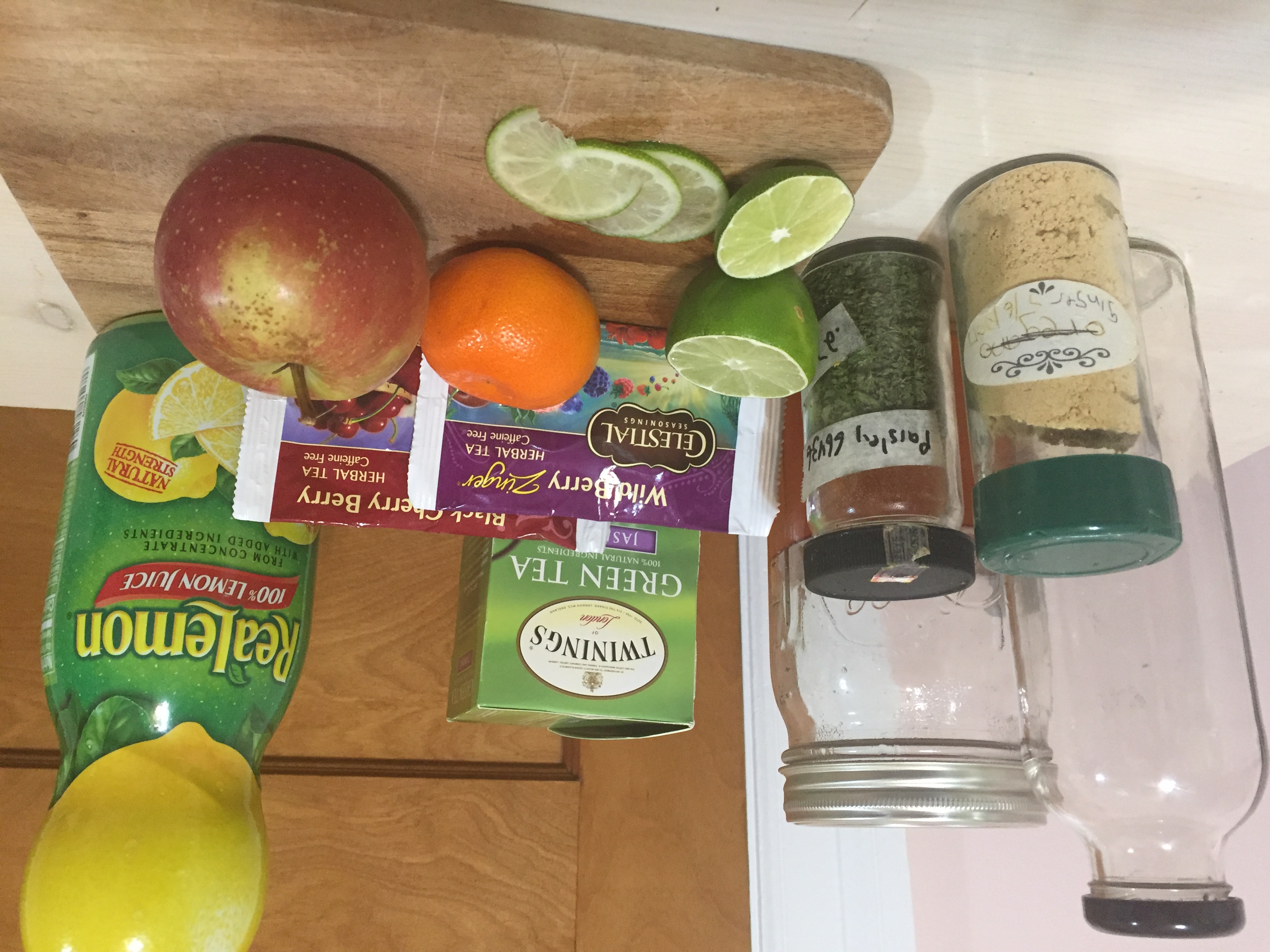Some of the recommended materials listed above, gathered on a kitchen counter. In the back we see a glass bottle and jar with lids, various packs of tea bags, and a bottle of lemon juice. In the front there are a couple jars of spices and a cuttingboard holding an orange, and apple, and a partially sliced lime.
