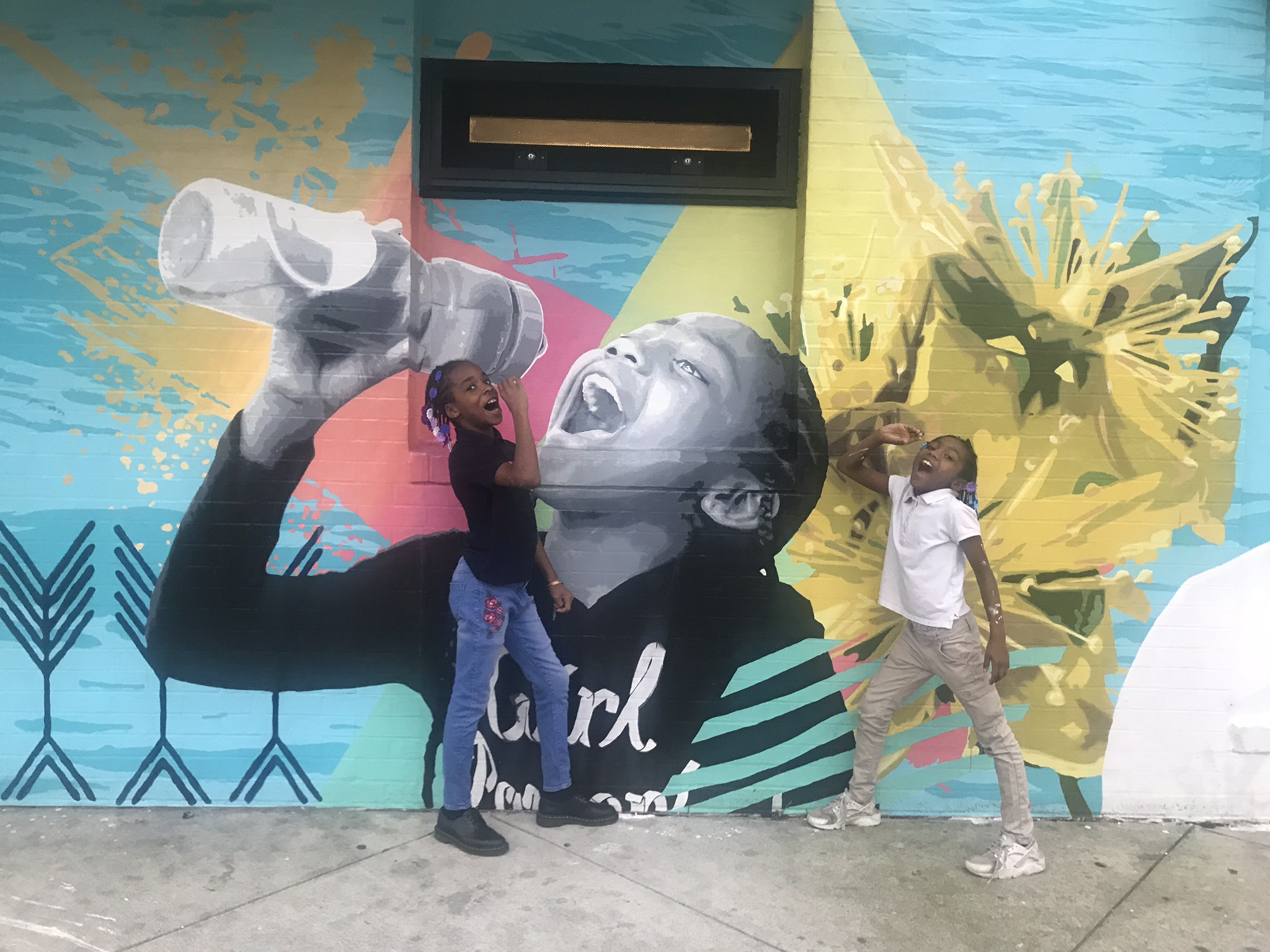The mural show a young girl with dark skin and braids drinking from a reusable water bottle, painted in shades of grey with colorful abstract designs on a light blue background that looks like water. The model and another young girl stand in front of the wall mimicing her pose