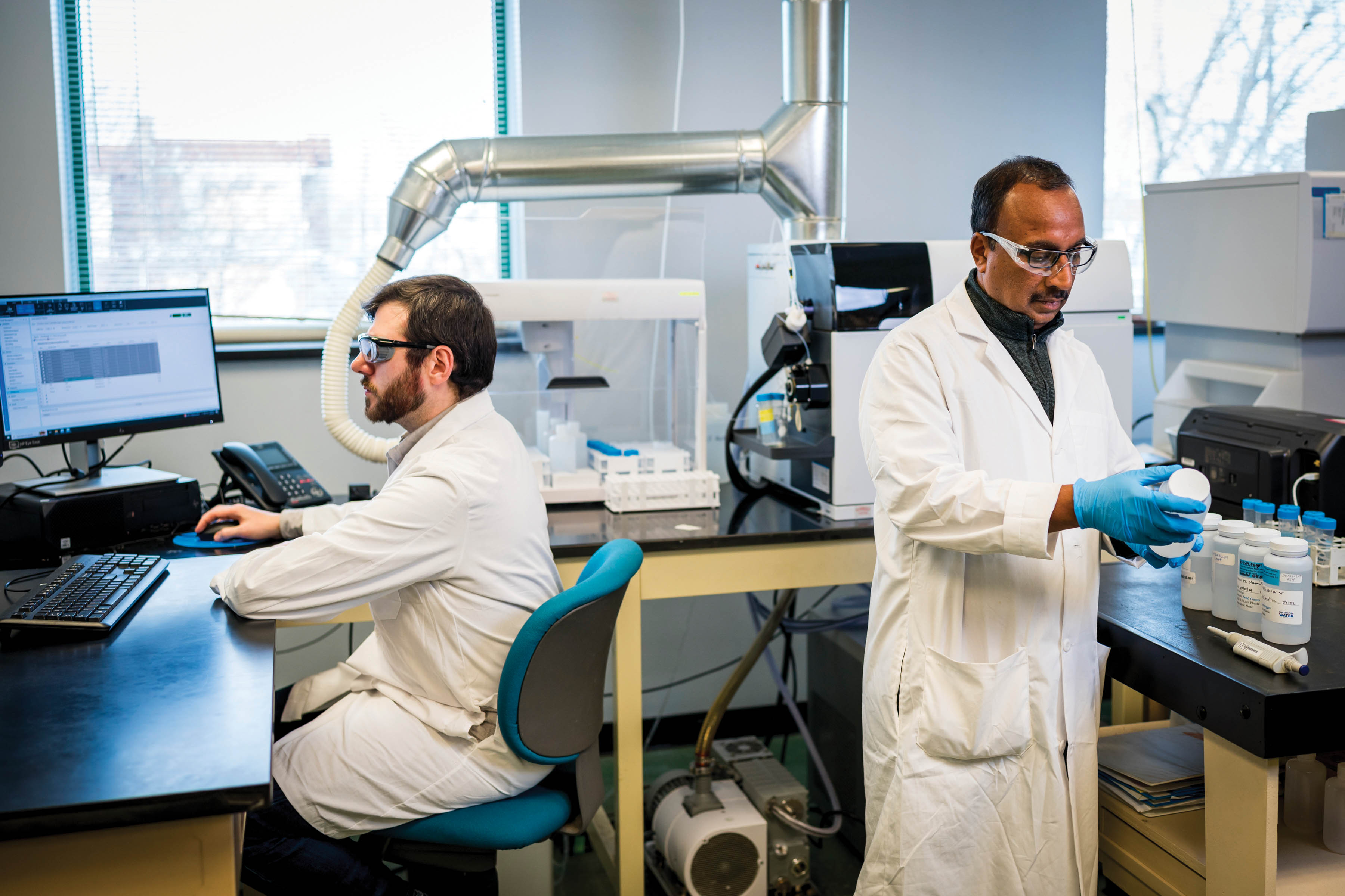 Two scientists, wearing white lab coats and safety glasses, at work in one of our labs. The one on the left is sitting at a lab table, using a computer. The one on the right is standing at another lab table, wearing blue disposable gloves, examining a water sample collection bottle, with several others lined up on the table. Various machines and a window are visible in the background.