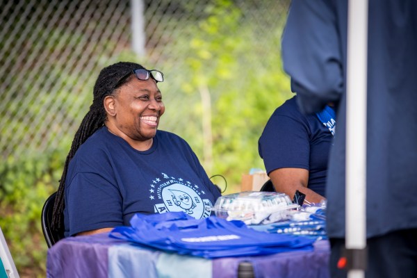 A PWD staff member with long braids tied back, wearing a blue PWD "At your service" t-shirt" sits behind a table at an open house with a huge smile.