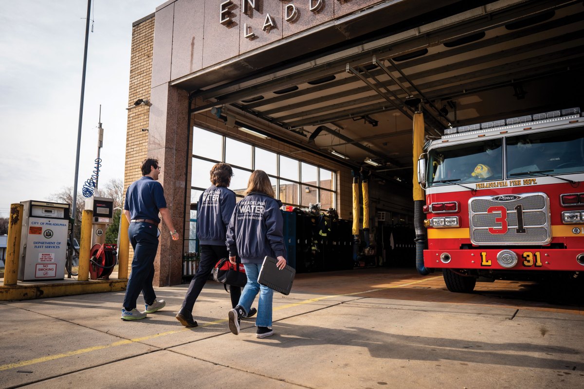 Three people, two of whom are wearing jackets with the Philadelphia Water Department logo on the back, are seen from behind walking into a fire station through an open double garage bay door with a fire engine parked on the right side.