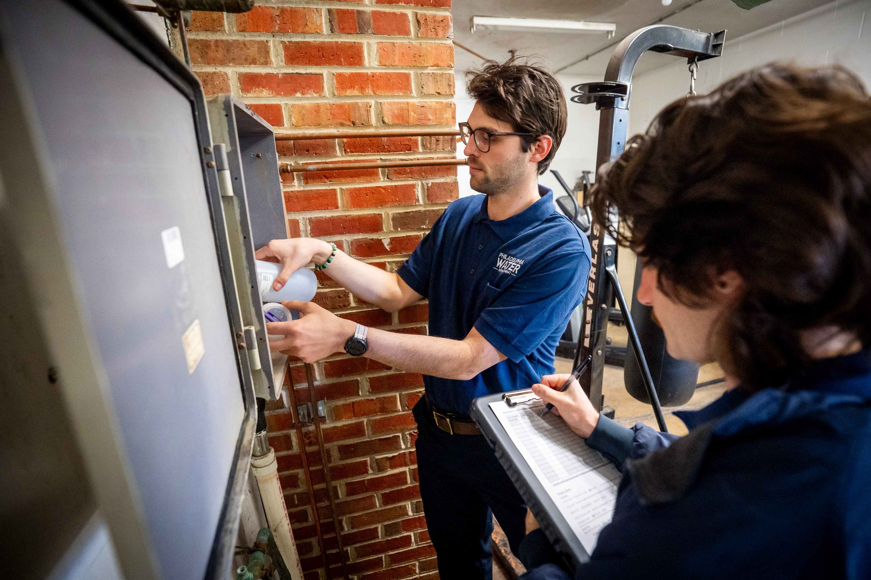 2 laboratory staff members collecting water samples in a fire station. One, wearing a blue PWD polo shirt, fills a sample bottle from a test tap in a gray utility box on a wall, while the other, back to the camera, makes notes on clipboard.