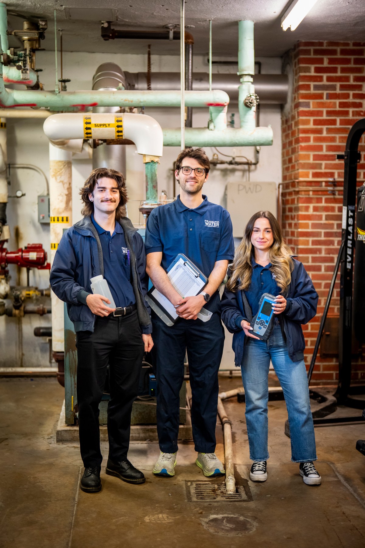 Three laboratory staff members pause to smile for the camera in the basement of a fire station. They are holding various tools used for collecting water samples.