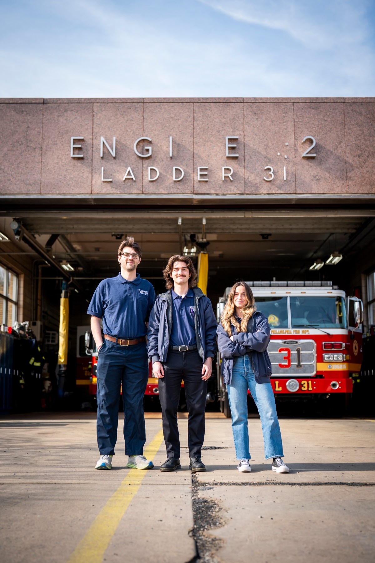 Three PWD BLS staff members pose in front of a fire station the Far Northeast section of Philadelphia. The bay door is open and you can see two firetrucks inside.