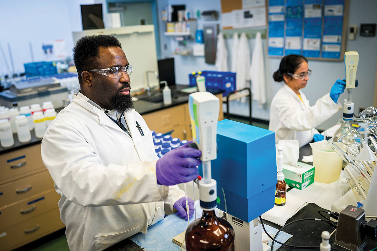 Two scientists, one with short textured hair and goatee, the other with long dark hair pulled back into a low bun, both wearing safety glasses, white lab coats, and disposable gloves, operate automatic titration machines in one of our labs. A variety of equipment and row of lab coats hanging on pegs on the wall are visible out of focus in the background.