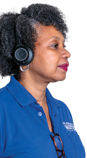 A contact center representative with tightly curled salt-and-pepper hair cut in a bob, wearing a blue polo with the PWD logo above a chest pocket and tortoise-shell glasses hanging from the neck, raspberry-colored lipstick, and silver hoop earrings peeking out from behind a black headset, looks off to the right with a serene expression.